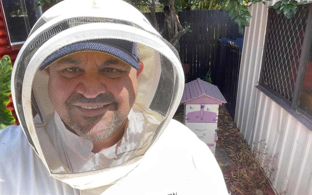 Man in bee suit in front of beehive.