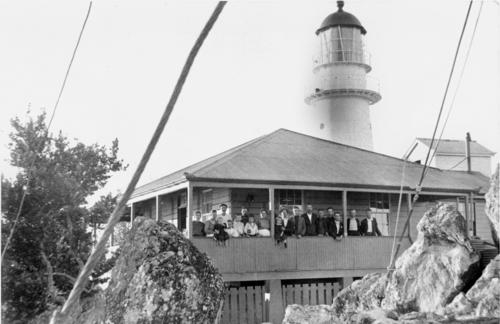 Black and white photo of Pine Islet's historic lighthouse with people in a building in the foreground.