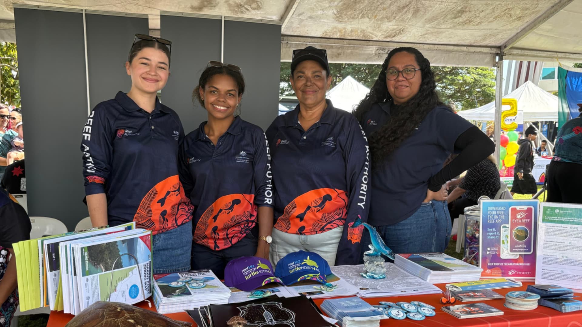 Four First Nations women in Reef Authority uniforms stand behind a Deadly Day Out stall.