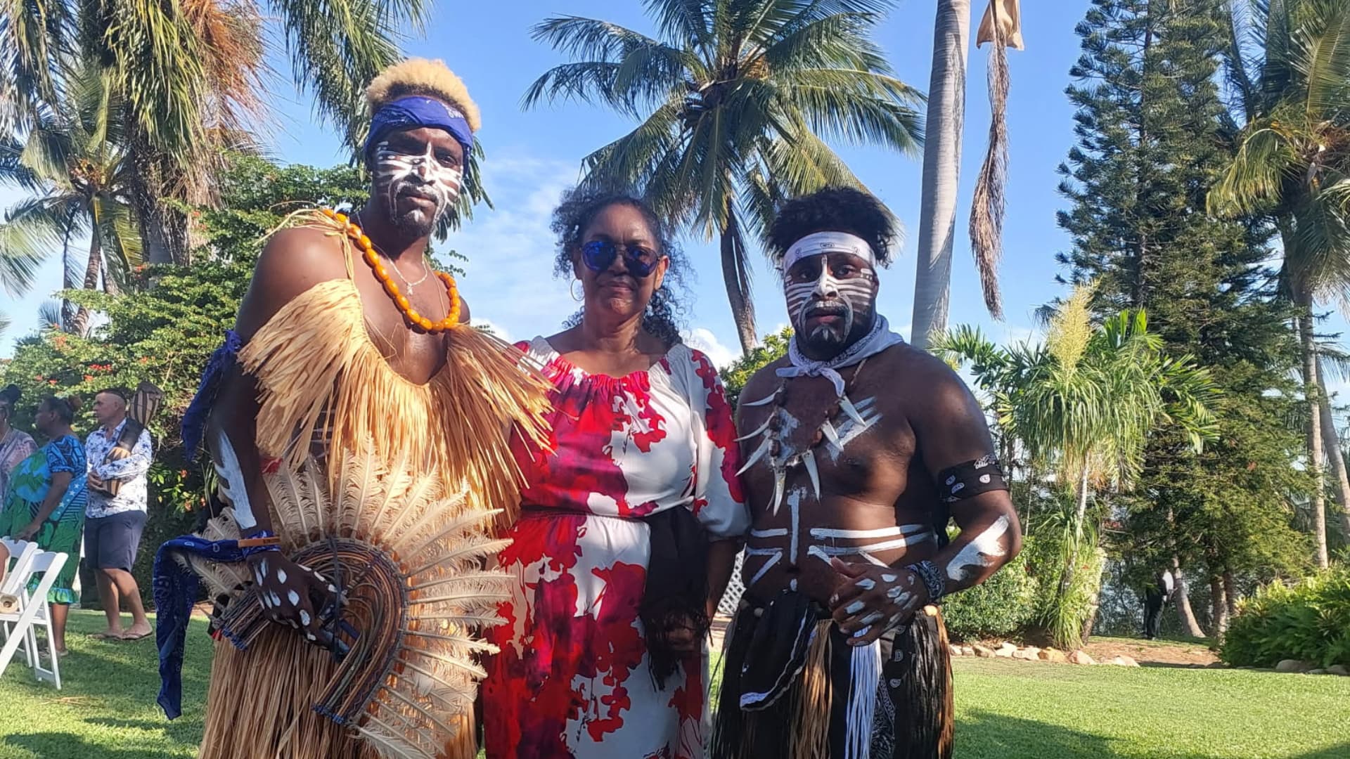 Marcia stands in between two men dressed in Indigenous traditional ceremonial gear.