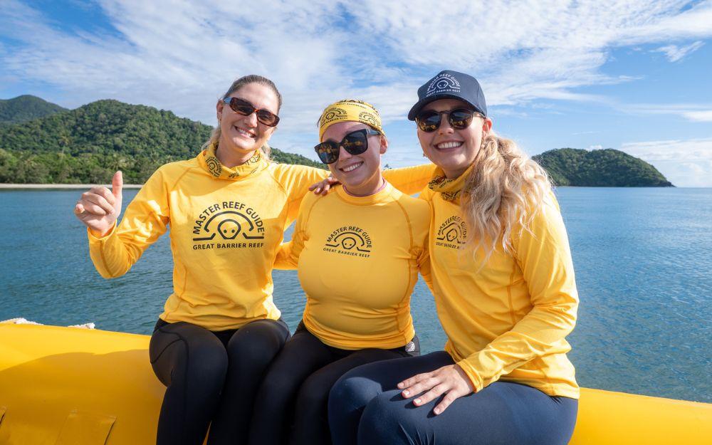 Three women in yellow shirts sit on the edge of a boat.