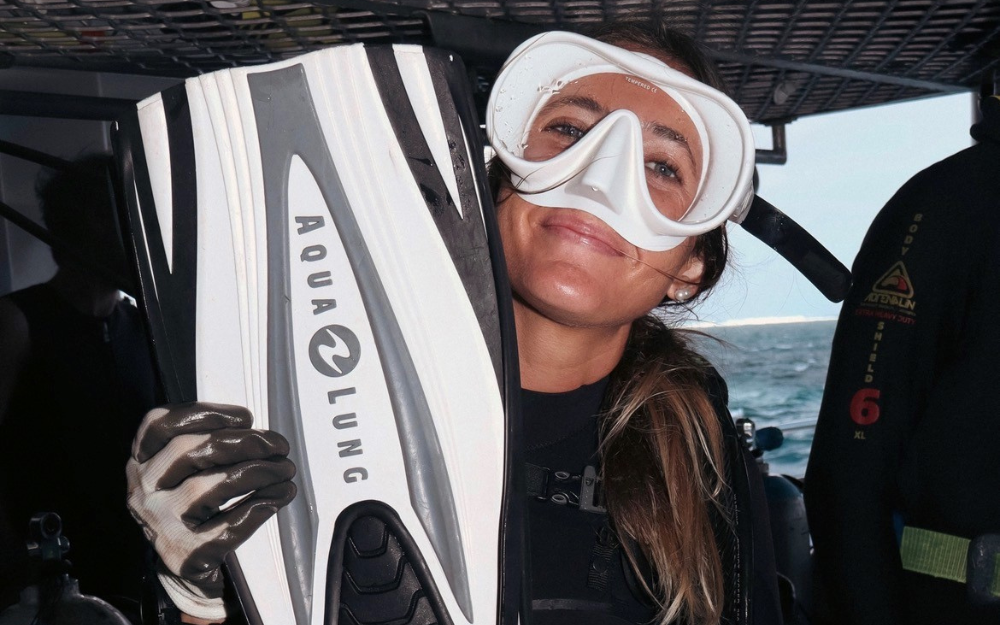 A woman in a snorkel mask holds her fins and smiles for the camera